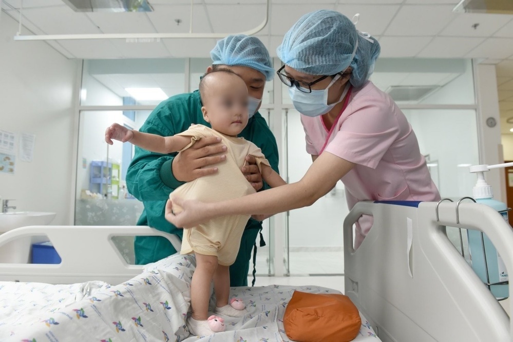 
Một trong 2 bé được tập đứng trên giường bệnh với sự giúp đỡ của bác sĩ (Ảnh: BVCC)